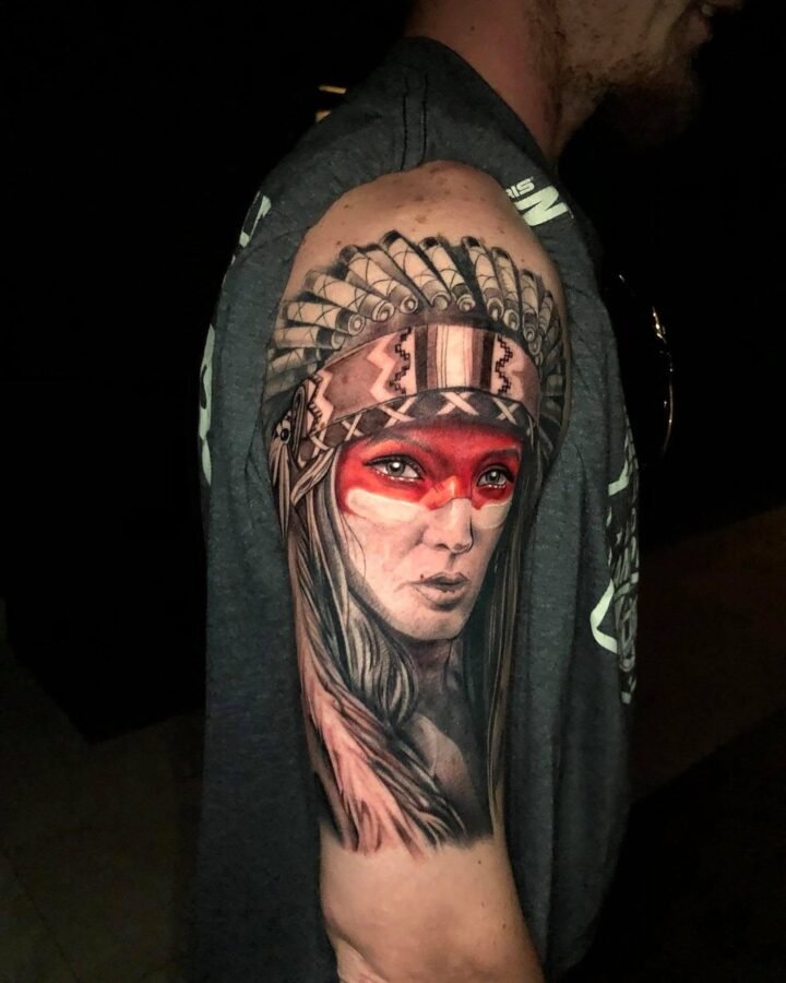 Portrait tattoo of a woman in a feathered headdress with red face paint on the upper arm