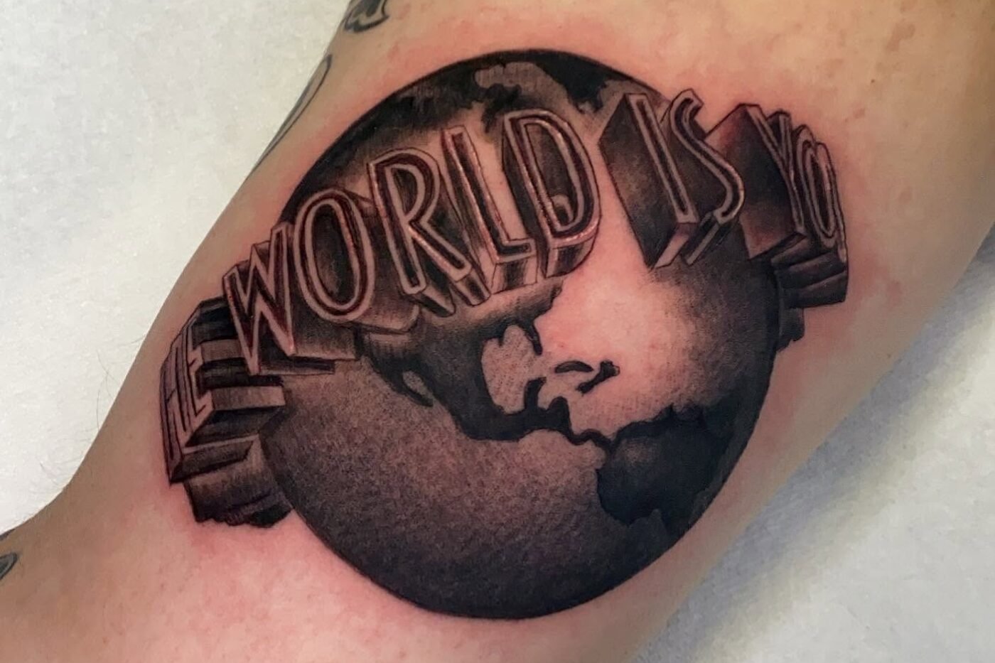 Black and grey tattoo of a globe reading The World Is Yours on the inner arm