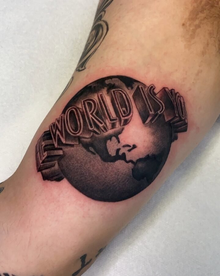 Black and grey tattoo of a globe reading The World Is Yours on the inner arm